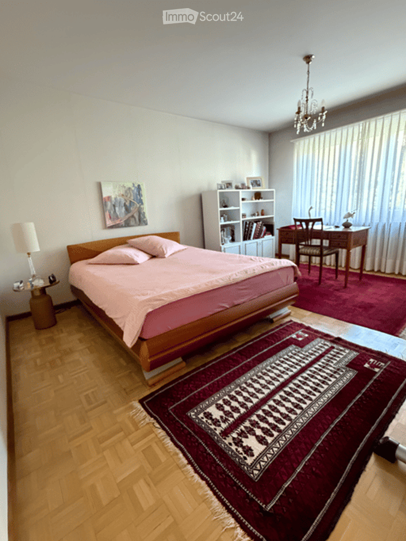 Spacious bedroom with wooden flooring, a large bed with pink bedding, a nightstand with a lamp, a desk with a chair, a bookshelf, and a decorative rug.