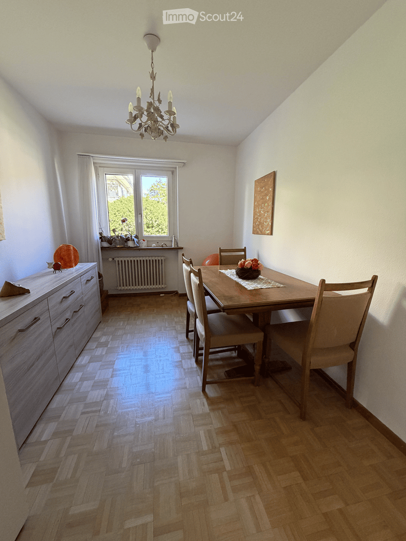 Dining room with wooden herringbone flooring, a chandelier, a dining table with chairs, a cabinet, and a window with white curtains.