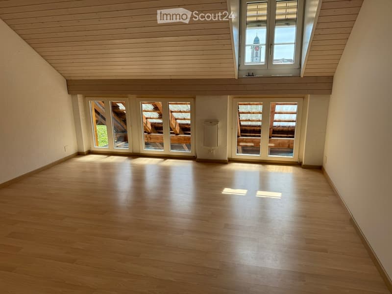 Empty room with wooden flooring and large windows, allowing natural light to fill the space. The walls are painted a light color, and the ceiling has wooden paneling.