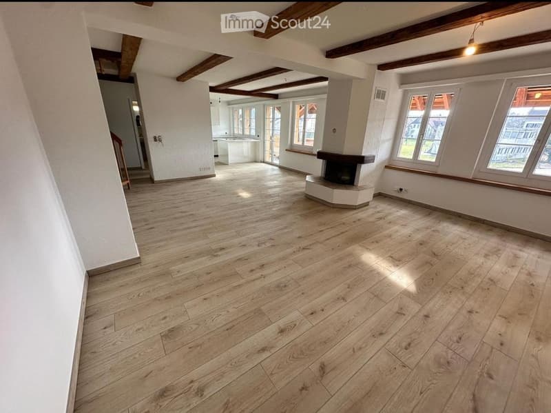 Empty room with wooden flooring, white walls, exposed wooden beams on the ceiling, and a fireplace. Large windows allow natural light to enter.