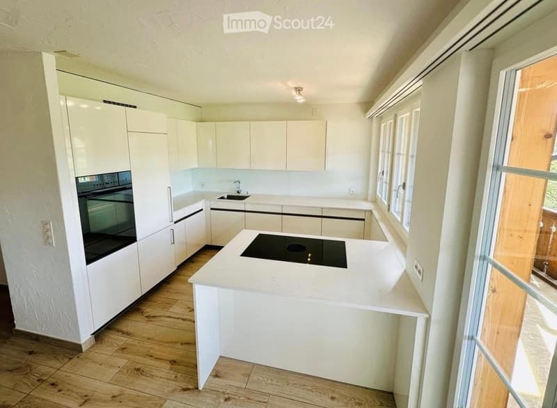 Modern kitchen with white cabinets, an island with an induction cooktop, and a built-in oven. The space features wooden flooring and large windows allowing natural light.
