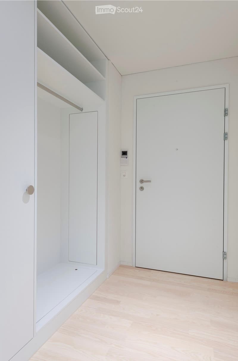 Empty room with light wooden flooring, white walls, built-in shelving unit, and a closed white door.