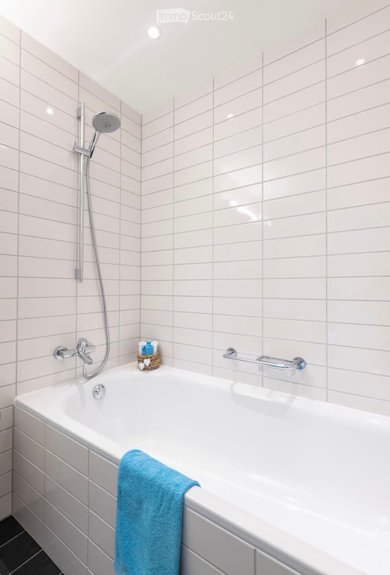 Modern bathroom with a white bathtub, tiled walls, a showerhead, a towel, and a handrail.