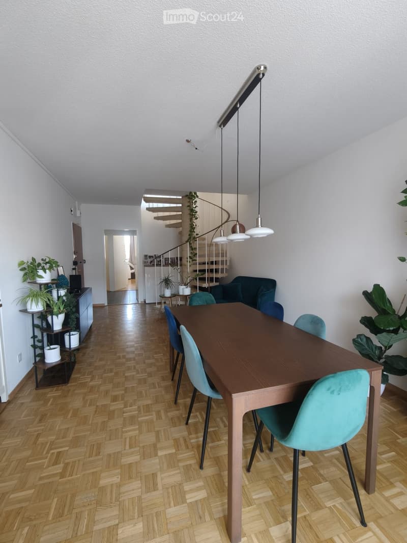 Dining area with a wooden dining table, teal chairs, a dark blue sofa, and several potted plants. A staircase leads to another level, and pendant lights hang from the ceiling.