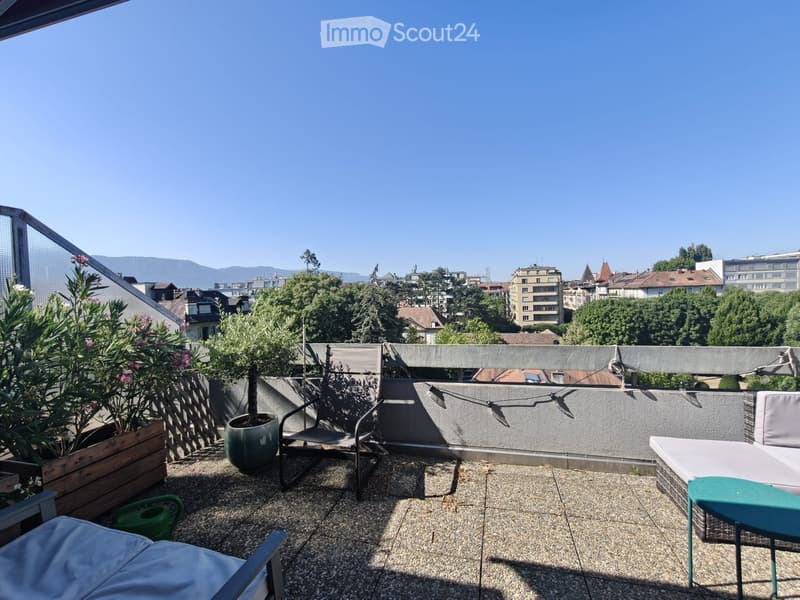 A rooftop terrace with a view of the city and mountains, featuring a sofa, chairs, a small table, and potted plants.
