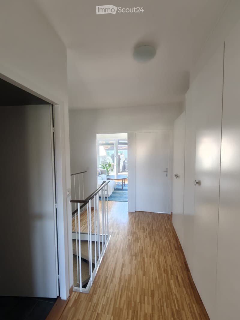 Bright hallway with wooden flooring, white walls, and a staircase leading to another level. A door on the right leads to a room with visible furniture.