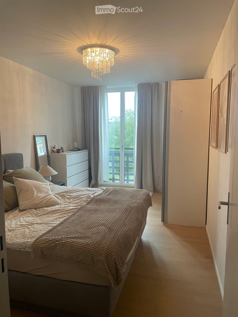 Well-lit bedroom with a large bed, nightstand, mirror, and a chandelier light fixture. The room features a large window with curtains and a view of a balcony.