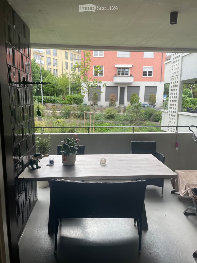 Balcony with a wooden table, chairs, and a potted plant, overlooking a residential area with greenery and buildings.