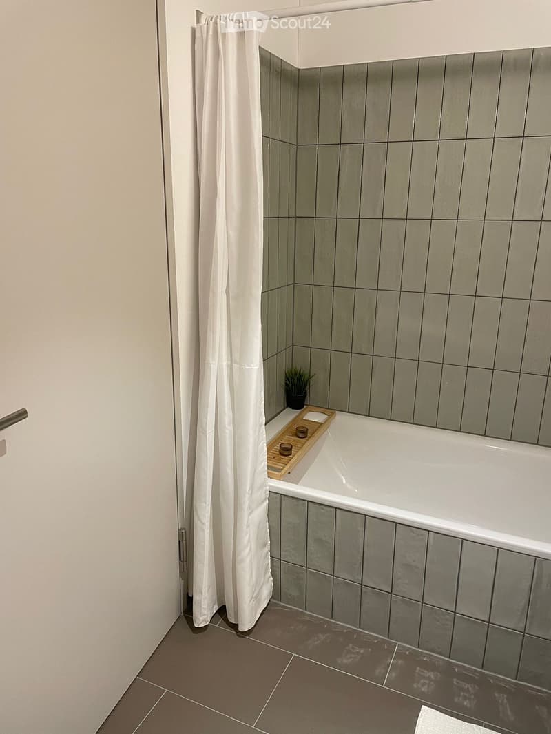 Modern bathroom with a bathtub, shower curtain, wooden tray with cups, and a small plant on the edge of the tub.
