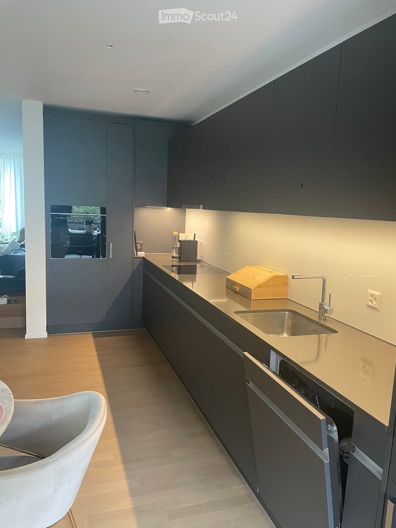 Modern kitchen with dark grey cabinets, light wooden countertop, stainless steel sink, and a white chair visible in the foreground.