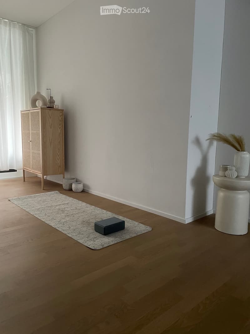 Minimalist room with light wooden flooring, white walls, a small wooden cabinet with decorative items, a white stool with vases, and a grey rug with a dark block on it.