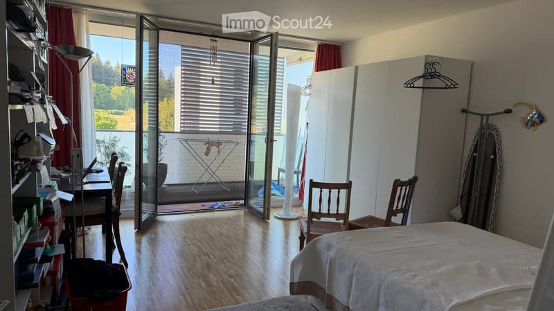 Bedroom with wooden flooring, large glass balcony door, bed, chairs, table, lamp, wardrobe, and ironing board visible.