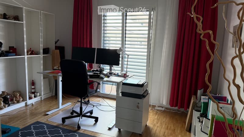 A well-organized home office with a desk, two monitors, a chair, a lamp, shelves with books and decorative items, and a printer on a stand.