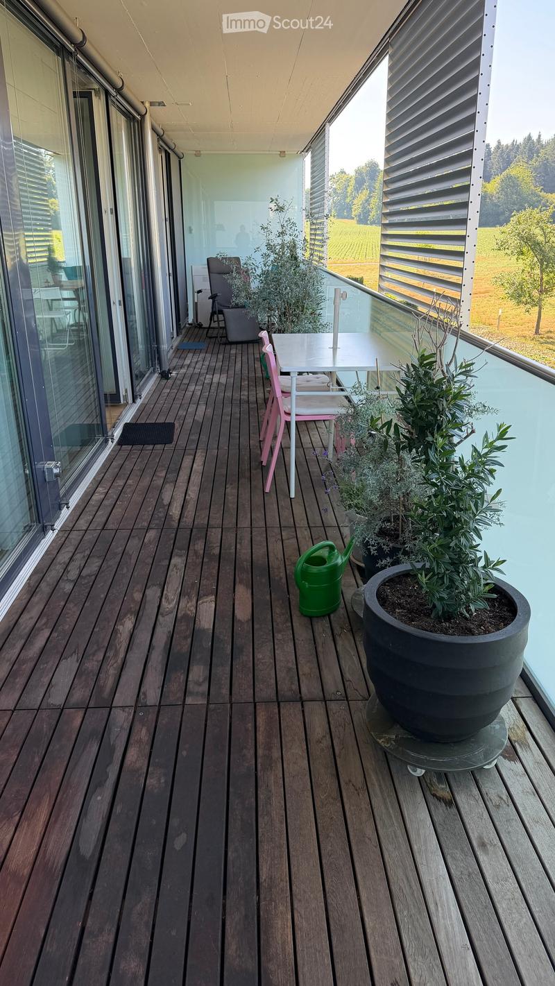 Wooden deck with a small table, pink chairs, potted plants, and a watering can, overlooking a scenic countryside view.