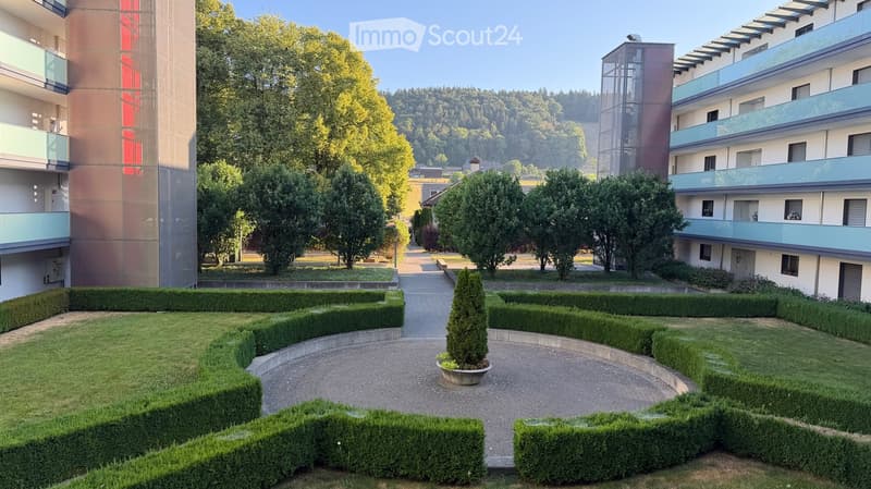 Well-maintained garden area with neatly trimmed hedges, a central tree, and a paved walkway leading to modern apartment buildings.