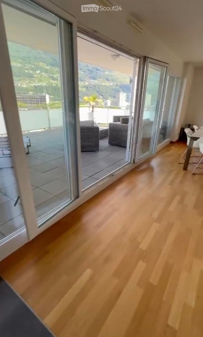 Bright living room with wooden flooring, large glass doors leading to a balcony with outdoor seating and scenic mountain view.