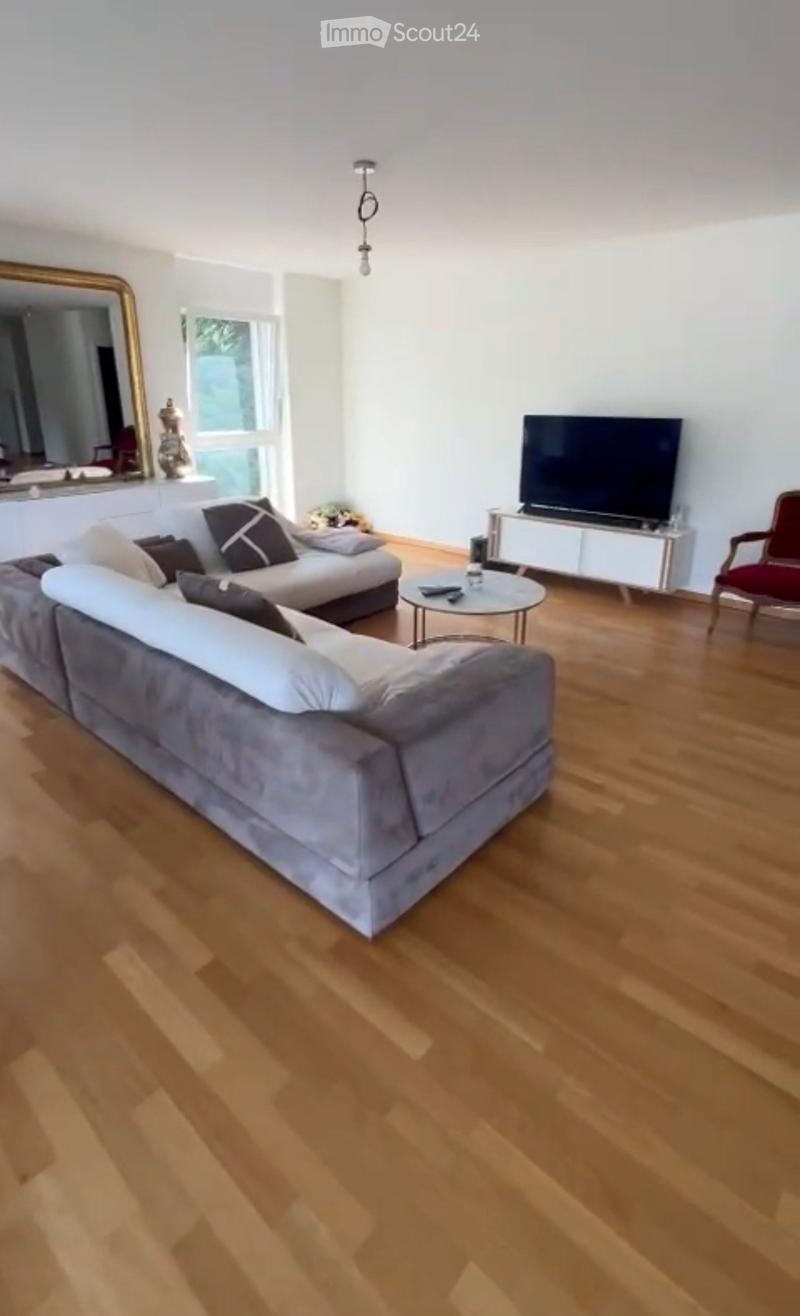 Modern living room with light wooden flooring, a grey sectional sofa, a round coffee table, a red armchair, and a flat-screen TV on a white stand.