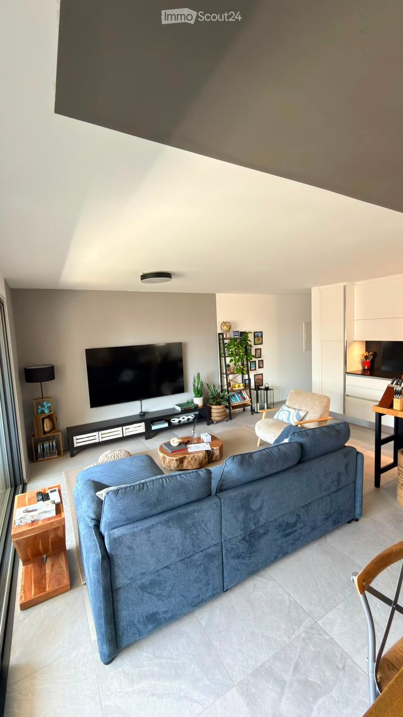 Modern living room with a blue sectional sofa, coffee table, armchair, TV, shelves, and plants.