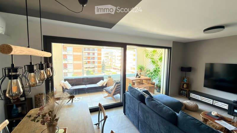 Modern living room with large sliding glass doors leading to a balcony, blue sofa, wooden dining table, hanging pendant lights, and a TV stand.