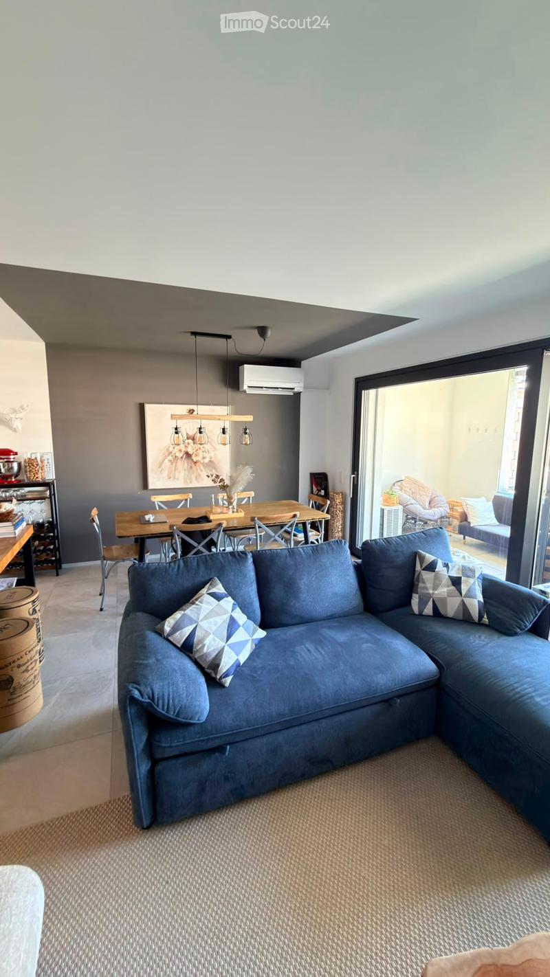 Modern living room with a blue sectional sofa, dining table with chairs, bookshelf, and large windows leading to a balcony.