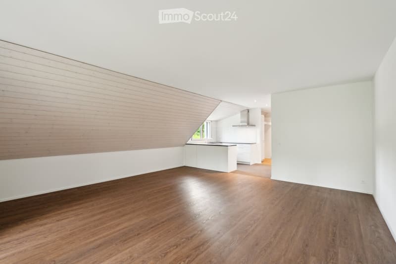 Empty room with wooden flooring, white walls, and a slanted wooden ceiling. The room is connected to a modern kitchen area with white cabinets and a stainless steel range hood.