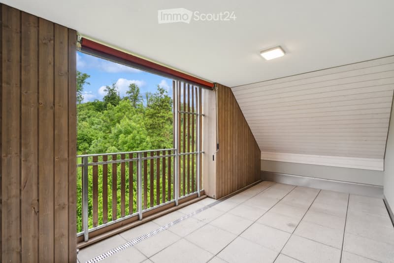 Modern balcony with wooden railings, tiled floor, and large glass window offering a view of green trees.