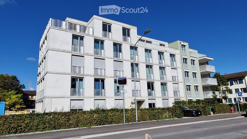 Modern multi-story residential building with balconies and large windows, surrounded by greenery and a parking area.