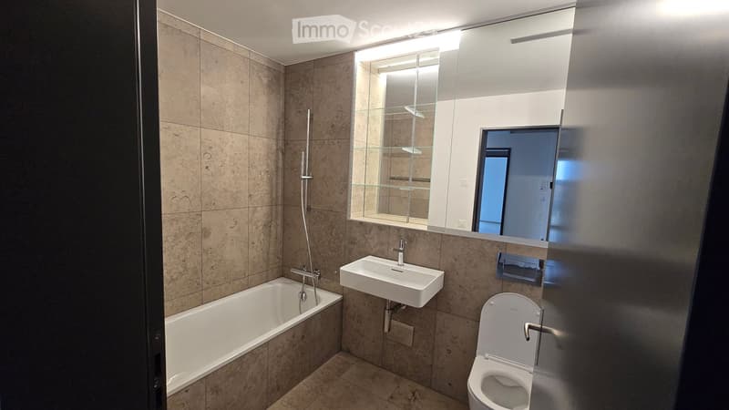Modern bathroom with beige tiled walls and floor, featuring a bathtub, sink, toilet, large mirror with built-in lighting, and a glass shelf.
