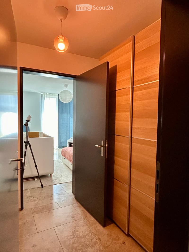 A narrow hallway with tiled flooring, warm lighting, and an open door leading to a bedroom with a bed and a tripod setup.