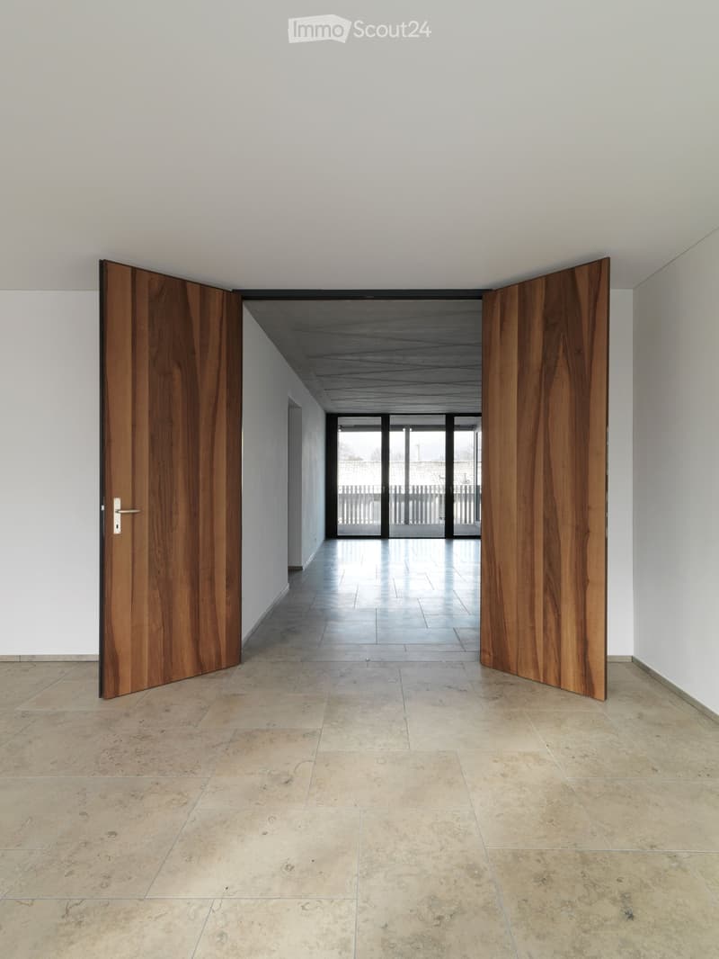 Empty hallway with light-colored tiled flooring, white walls, and large wooden double doors leading to an outdoor area with glass panels.