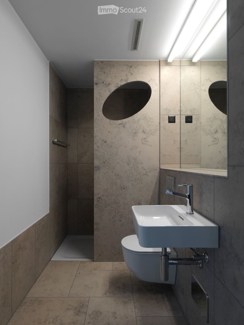 Modern bathroom with a sleek sink, chrome faucet, toilet, and large mirror with built-in lighting. The walls are tiled in a textured, neutral tone, and the floor is made of large, light brown tiles.
