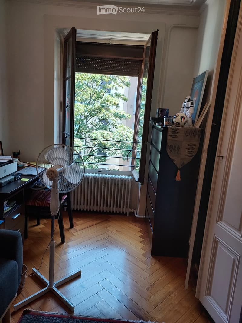 A furnished room with wooden flooring, a large open window leading to a balcony, a standing fan, a chair, a table with a laptop, and a refrigerator with various items on top.