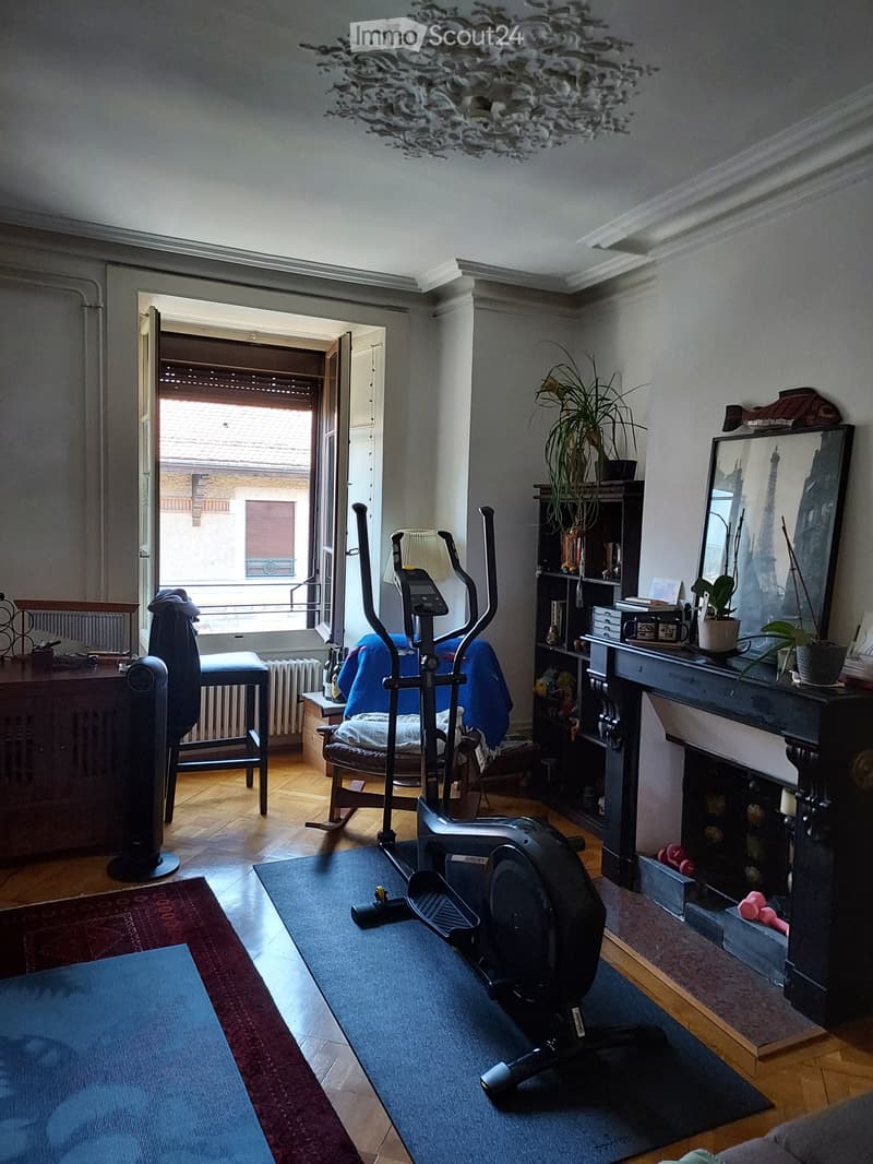 Living room with exercise bike, chair, table, mirror, plant, cabinet, fan, radiator, and a view of a balcony.