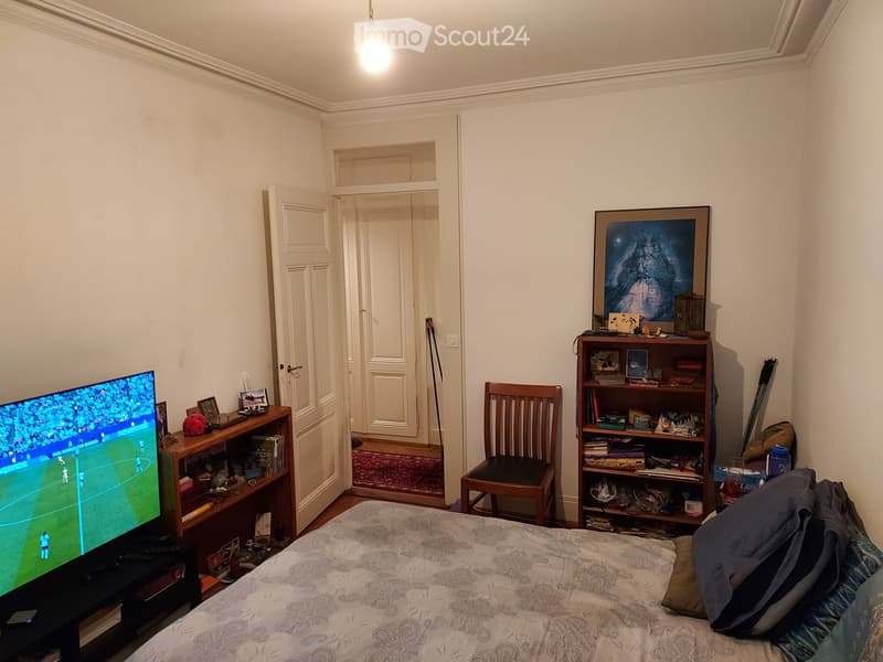 A furnished bedroom with a bed, a wooden chair, a shelf with books and decorative items, a TV screen, and a small table with a red ball and framed photos.