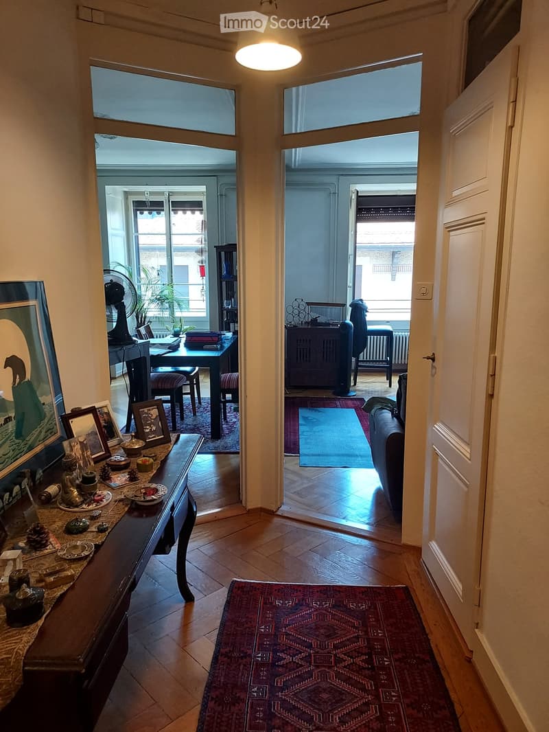 A well-lit hallway leads to a living area with a sofa, a coffee table, and a dining area with a table and chairs. The space features wooden flooring, decorative rugs, and various decorative items on a side table.