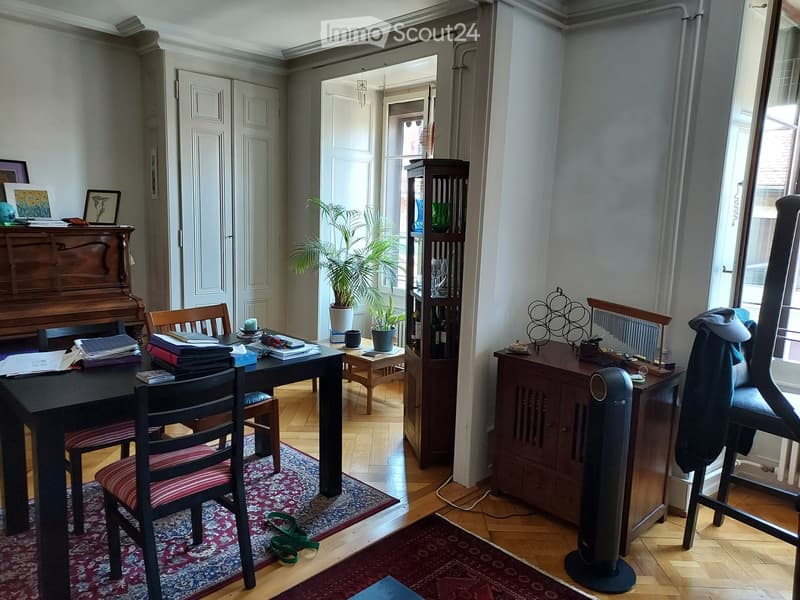 A well-furnished room with a wooden floor, a large table with chairs, a piano, a bookshelf, a fan, and a potted plant. The room has large windows and a door leading to another space.