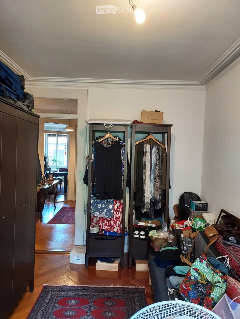 A furnished room with wooden flooring, a clothing rack holding various garments, a sofa with colorful cushions, a small table with a chair, and a cabinet. The room also contains a backpack and a handbag on the floor.