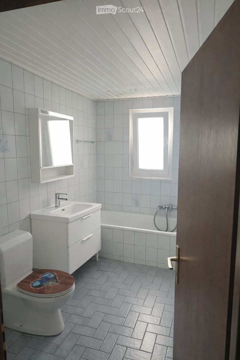 Modern bathroom with white tiles, a wooden toilet seat, a white sink with a mirror cabinet, and a bathtub. The room features a skylight window and grey tiled flooring.