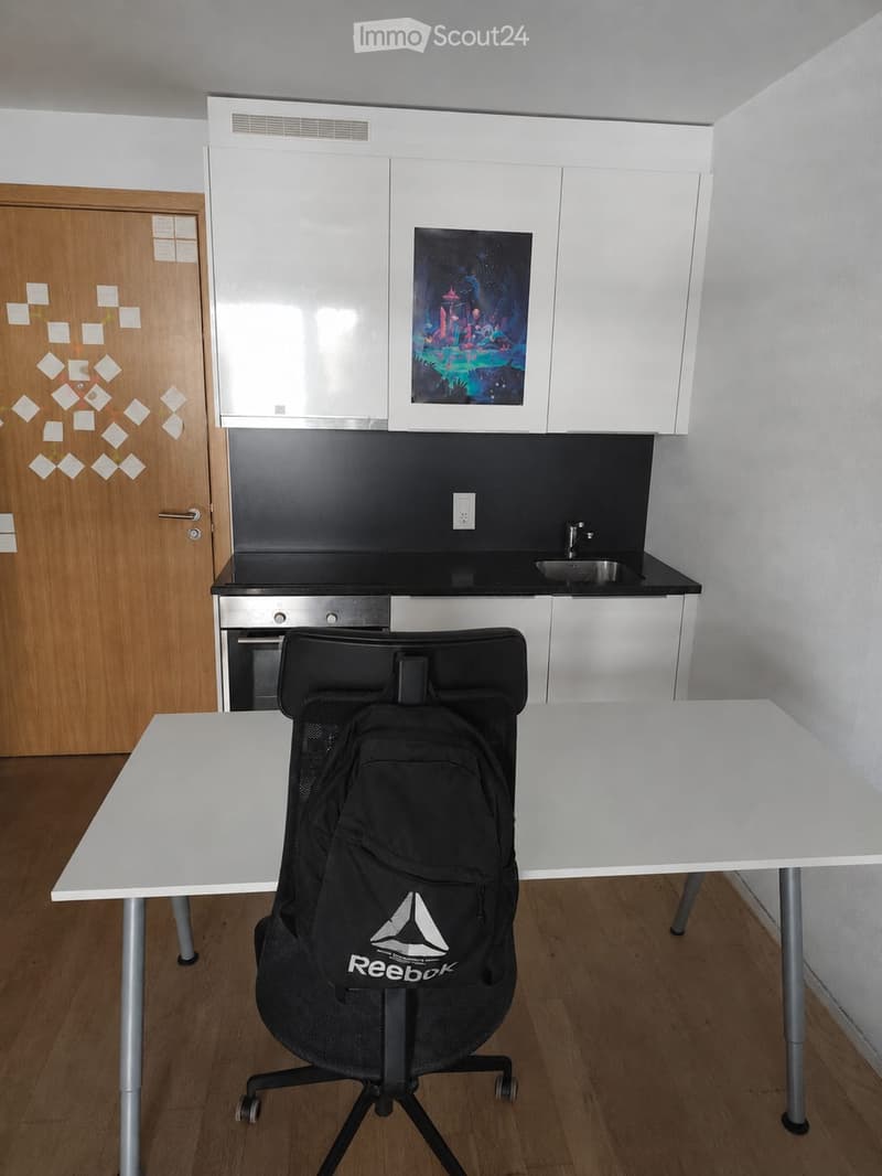 A modern, compact room with a white table, a black Reebok backpack, a black office chair, white cabinets, a sink, and a small oven. The room features a wooden floor and a colorful picture on the wall.