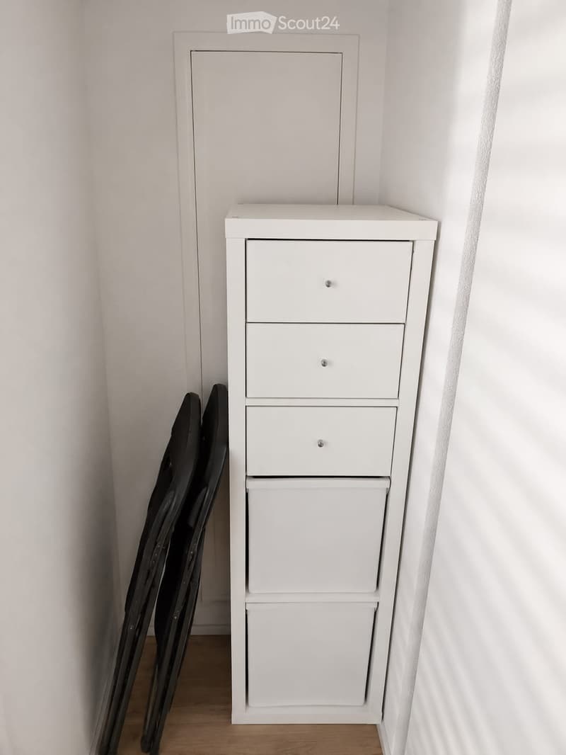 A small, unfurnished room with a white cabinet featuring multiple drawers and a folded black chair leaning against the wall.