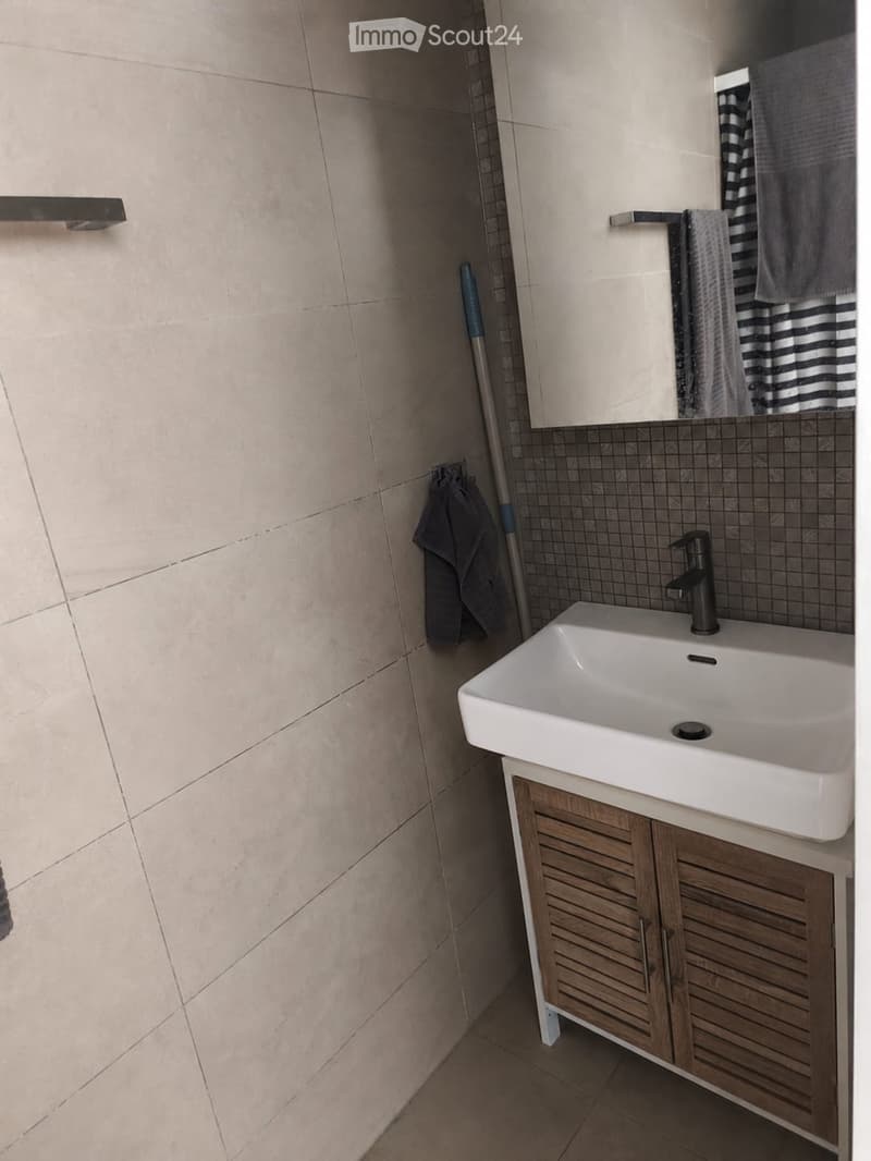 Modern bathroom with a white sink, wooden cabinet, and a mirror above it. Beige tiled walls and floor, with a towel and a mop visible.