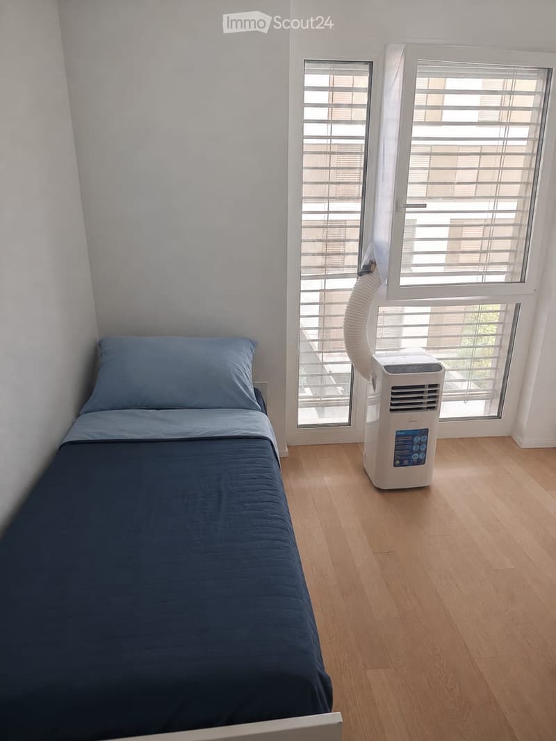 A small bedroom with a single bed, light blue bedding, and a portable air conditioner near the window.