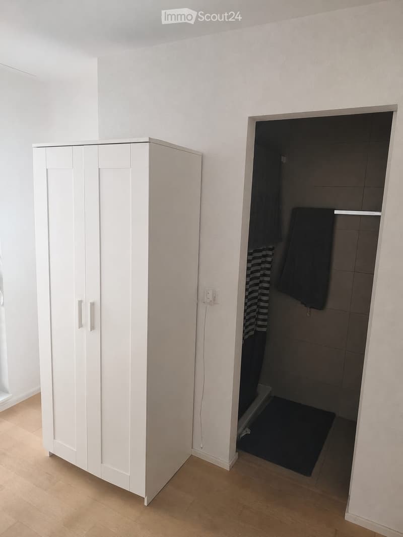 Empty room with white walls, light wooden flooring, and a white wardrobe. An adjacent bathroom is visible with dark tiles, towels, and a small rug.