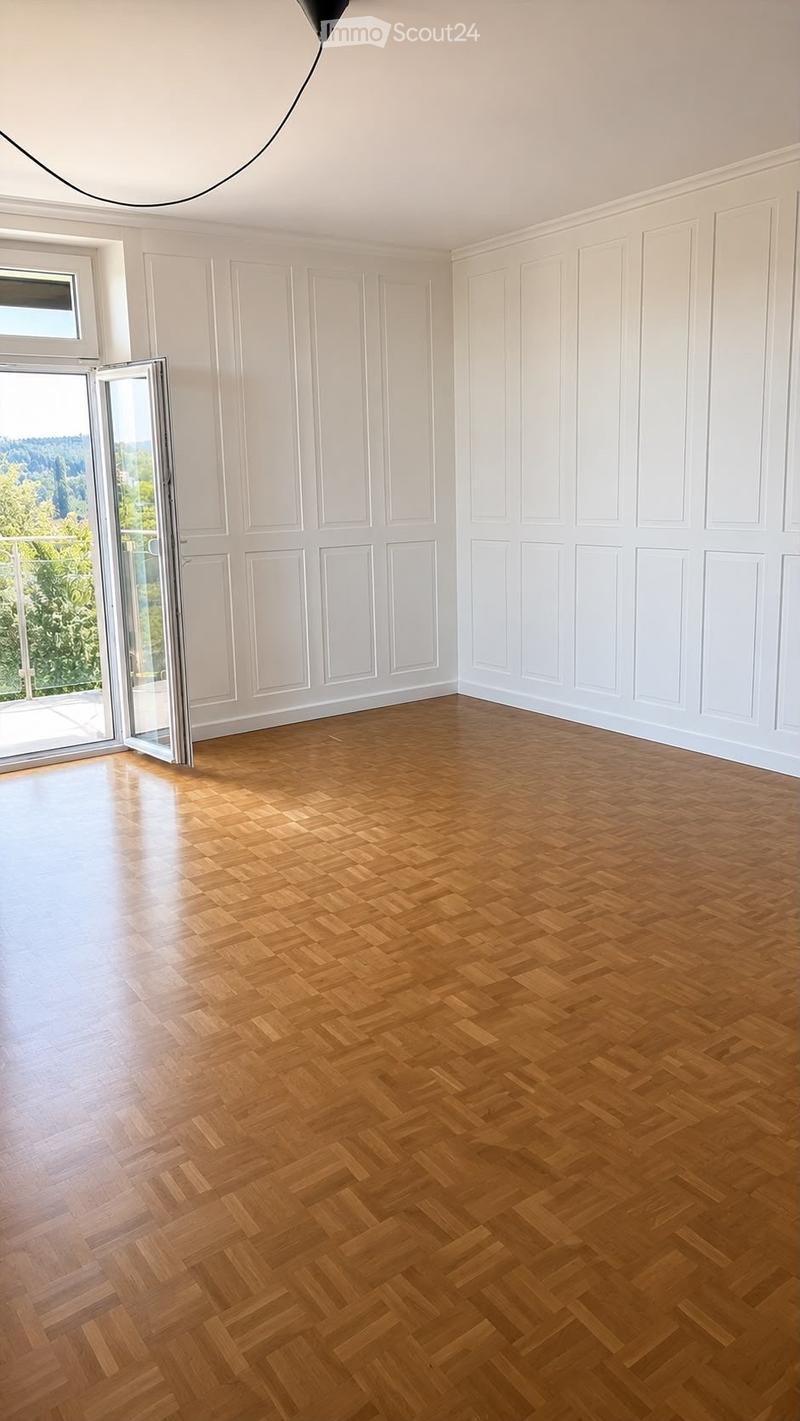 Empty room with wooden herringbone flooring, white paneled walls, large window, and sliding glass door leading to a balcony.