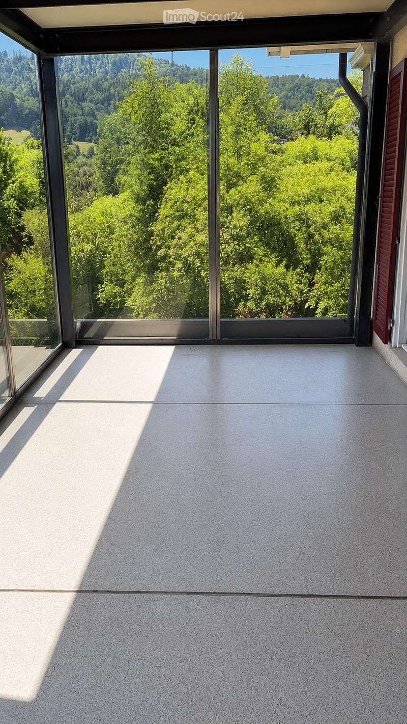 Modern balcony with large glass sliding doors, concrete floor, and scenic view of green trees and hills.