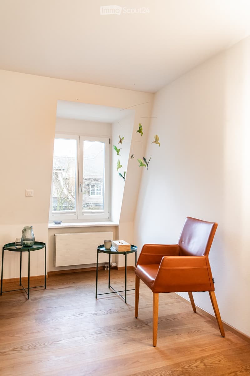 A small, well-lit room with wooden flooring, a large window, a brown leather chair, two small round tables, and decorative items on the tables.