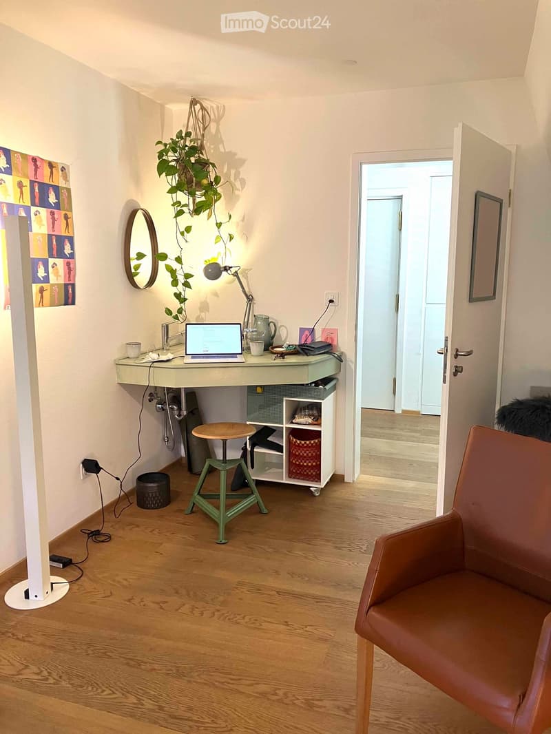 A modern, well-lit room with a wooden floor, a small desk with a laptop, a green stool, a brown leather chair, a hanging plant, a mirror, and a framed picture on the wall.