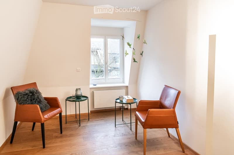 A cozy living room with light-colored walls, wooden flooring, and two leather chairs flanking a small round table. A window with a view of the outside is visible, and decorative items like a vase and a pillow add warmth to the space.