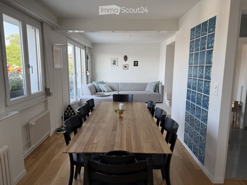 Bright living and dining area with a grey sofa, wooden dining table, black chairs, and decorative candle. Large windows and sliding glass doors lead to a garden view.