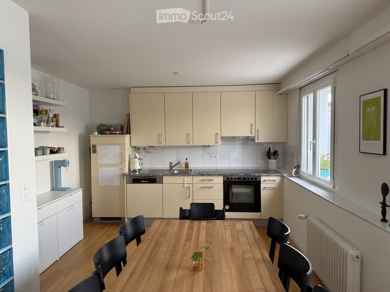 Modern kitchen with light-colored cabinets, a central island, a wooden dining table with black chairs, and various appliances including a coffee maker and a framed picture on the wall.